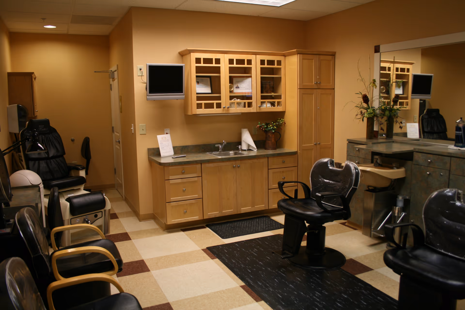 Interior view of a salon area in a senior living facility featuring multiple black salon chairs, a sink for hair washing, wooden cabinets with glass doors, a countertop with a sink, a wall-mounted TV, and a large mirror with a floral arrangement.