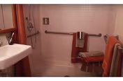 A bathroom with a walk-in shower featuring a grab bar and a foldable shower seat. The walls are tiled in light pink, and there are several towels hanging on bars, including a red and beige towel on the grab bar and orange towels on a rack. A white sink with a faucet is visible on the left side.