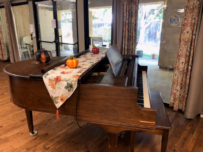 A wooden grand piano with a decorative autumn-themed table runner and small pumpkin decorations on top, situated in a room with wooden flooring, large windows, and patterned curtains. Outside the windows, a garden area is visible.