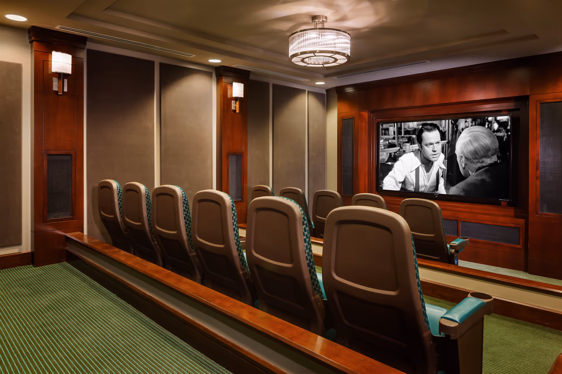 A small theater room with two rows of cushioned seats facing a large screen displaying a black and white movie scene. The room features wood paneling, wall-mounted lights, and a modern ceiling light fixture.