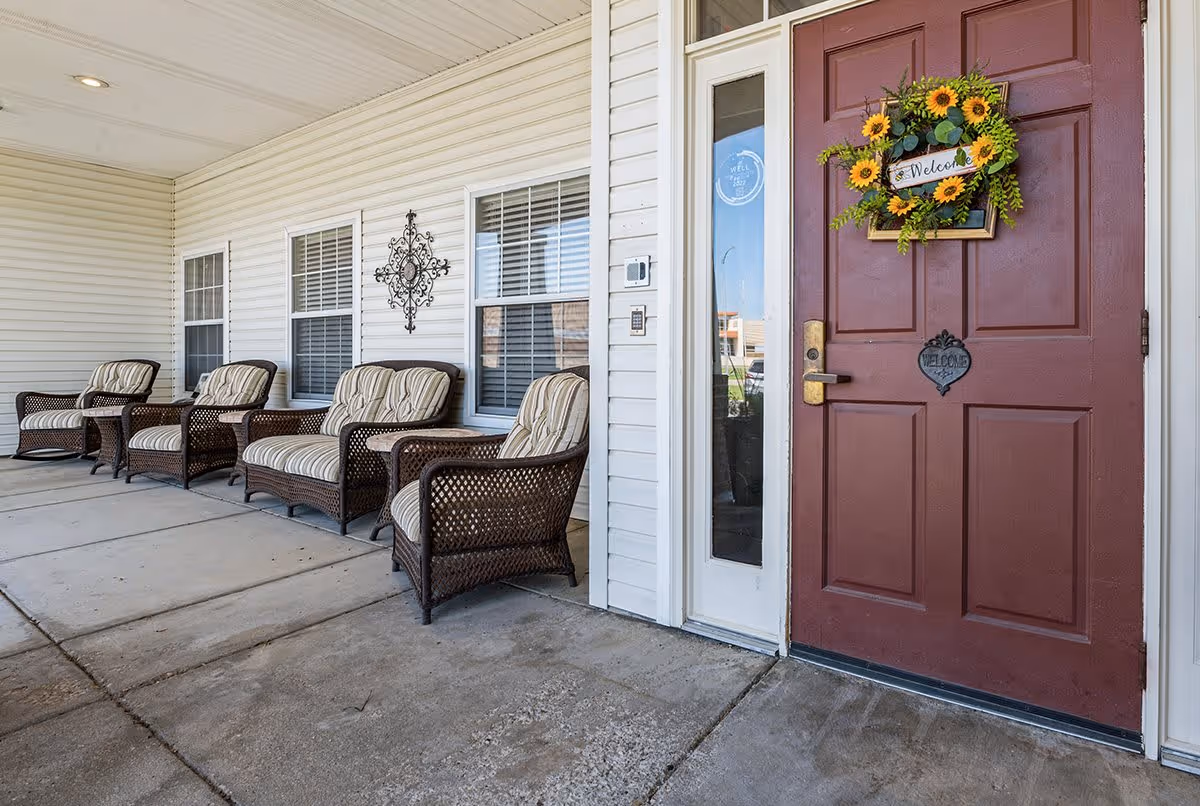 Covered outdoor porch area with four cushioned wicker chairs arranged along a white siding wall with three windows. A red door with a sunflower wreath and a welcome sign is visible on the right side.