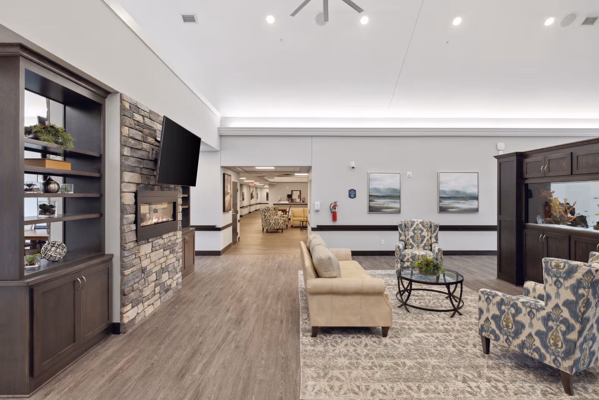 A spacious senior living facility common area featuring a beige sofa, two patterned armchairs, a glass coffee table with a plant, a large built-in aquarium, a stone fireplace with a mounted flat-screen TV above it, and wooden shelving units. The room has wood flooring, a patterned area rug, and neutral-colored walls with two landscape paintings. A hallway is visible in the background with additional seating and lighting.
