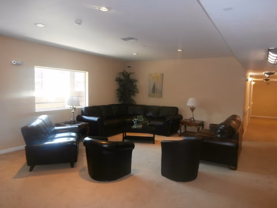 A spacious living room area with beige walls and carpet, featuring a black leather sectional sofa, two black leather armchairs, and a black leather loveseat. There are two wooden side tables with lamps on either side of the sectional, a small coffee table with a plant centerpiece, a large window letting in natural light, and a green potted plant in the corner. A hallway extends from the living room area.