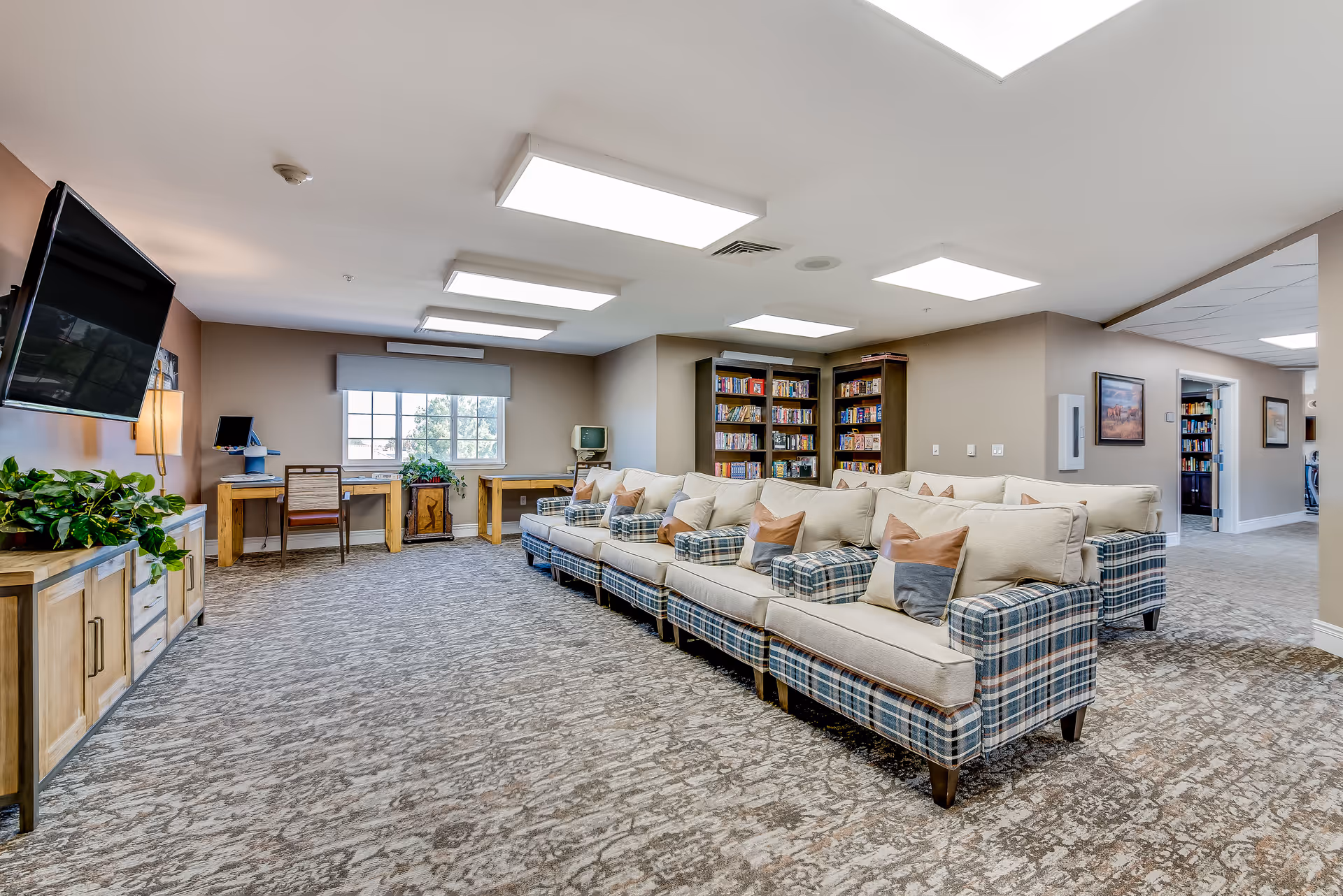 Spacious senior living lounge with a row of plaid sofas, a wall-mounted TV, bookshelves and desks by a window.