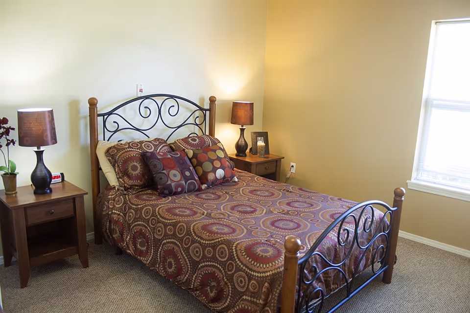 A bedroom with a double bed featuring a decorative metal and wood headboard and footboard. The bed is covered with a patterned bedspread and several matching pillows. There are two wooden nightstands on either side of the bed, each with a lamp and some decorative items. A window with blinds is on the right wall, allowing natural light into the room.