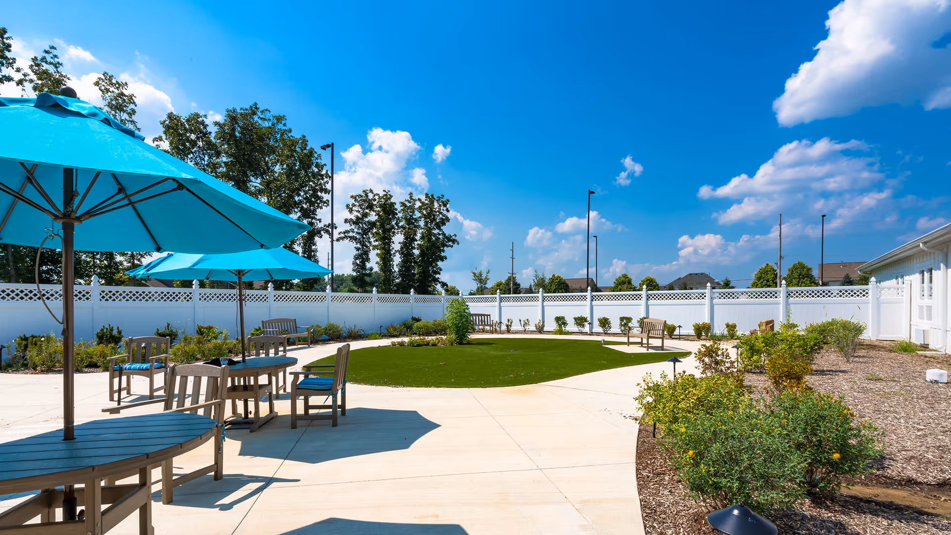 Outdoor patio area with several wooden tables and chairs under large turquoise umbrellas. The patio is surrounded by a white fence, with green shrubs and trees in the background under a bright blue sky with scattered clouds.