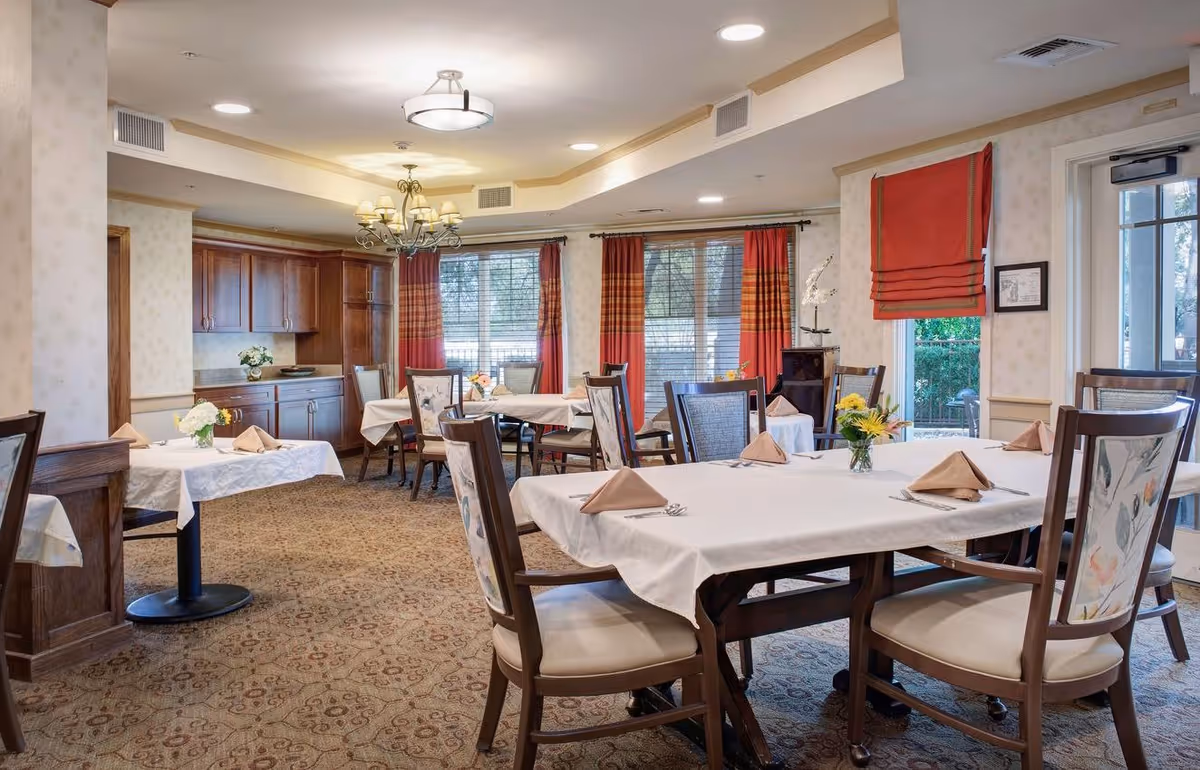A dining room in a senior living facility with several tables covered in white tablecloths, each set with folded napkins, silverware, and small flower arrangements. The room features patterned carpet, wooden chairs with floral upholstery, large windows with red and orange curtains, and a chandelier hanging from the ceiling. There is cabinetry along one wall and a door leading outside.