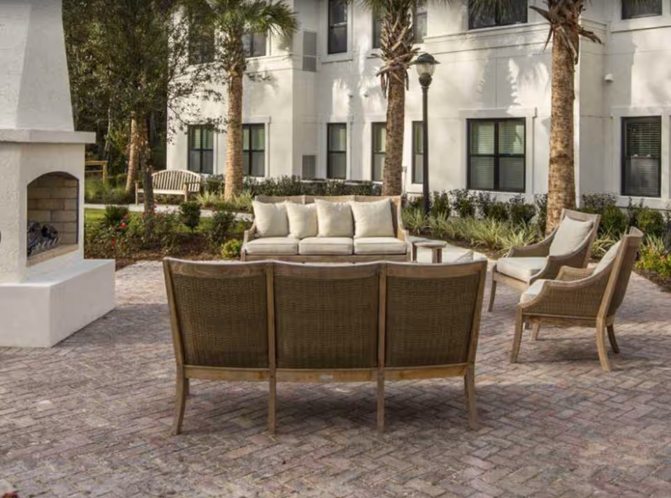 Outdoor patio area with cushioned seating including a sofa, chairs, and a bench, surrounded by palm trees and landscaping, adjacent to a white building with multiple windows. There is also a white outdoor fireplace on the left side.