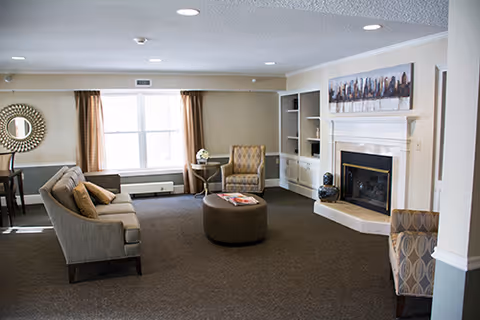 A spacious living room with a beige sofa, two patterned armchairs, a round ottoman in the center, a fireplace with a painting above it, built-in shelves, and a window with brown curtains letting in natural light.