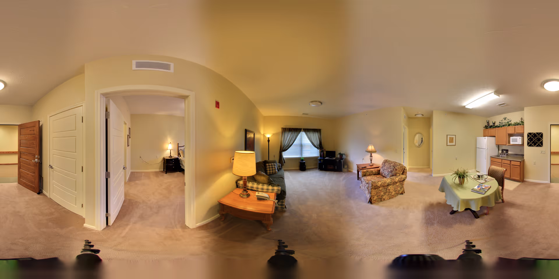 Panoramic view of a senior living facility apartment with a living room, dining area, kitchen, and a bedroom visible through an open door. The living room has a sofa, armchair, side tables with lamps, and a window with curtains. The dining area has a small table with a green tablecloth and two chairs. The kitchen includes a refrigerator, microwave, stove, and cabinets. The bedroom has a bed with a nightstand and lamp. The walls are painted light yellow and the floor is carpeted.