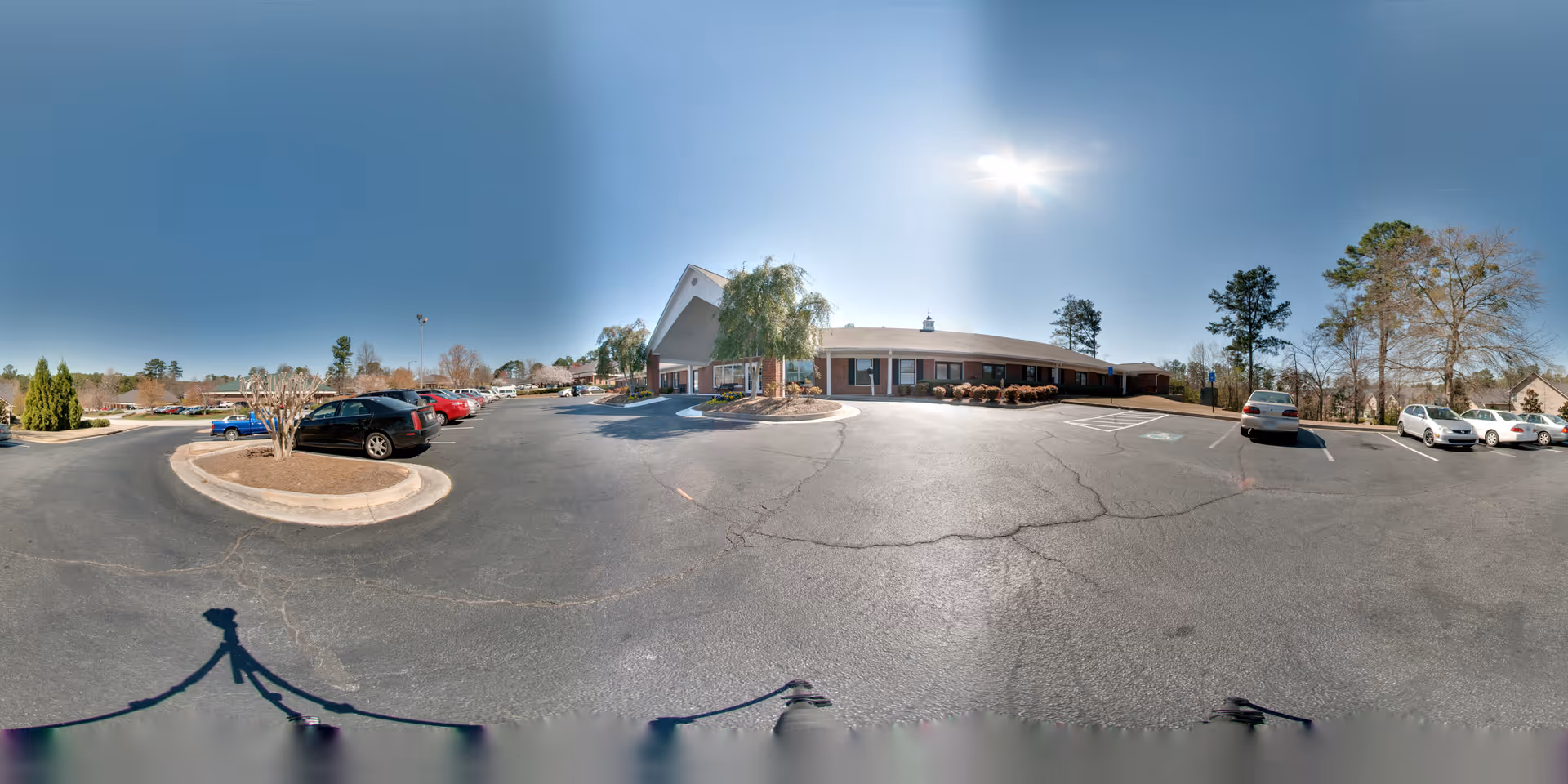 Wide panoramic view of the parking lot and front entrance of a single-story brick building under a clear blue sky with the sun shining brightly. Several cars are parked in the lot, and there are trees and shrubs around the building and parking area.