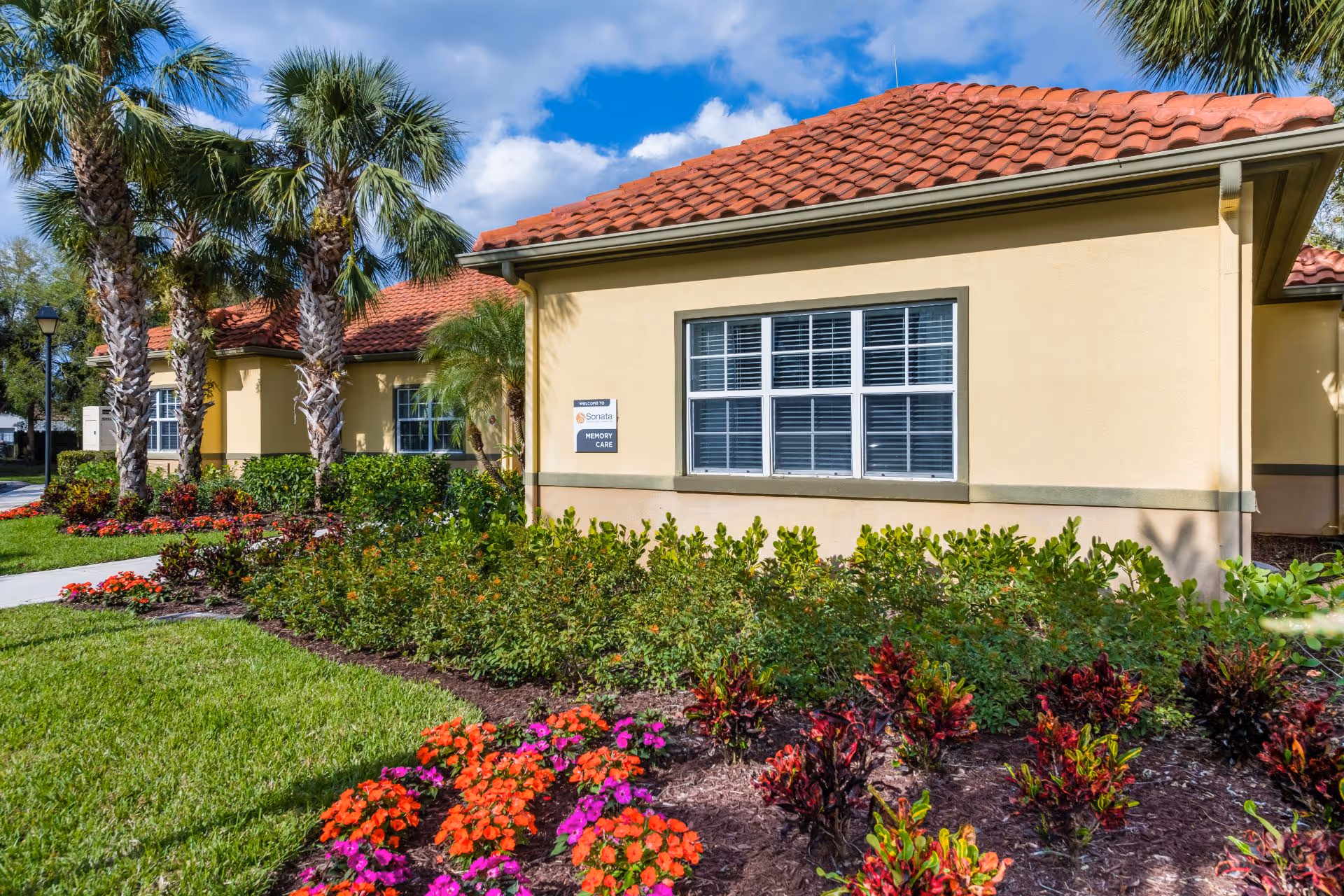 Exterior view of a single-story building with a red tile roof and beige walls, surrounded by landscaped gardens with colorful flowers and palm trees under a blue sky with some clouds. A sign on the building reads 'Sonata Memory Care'.