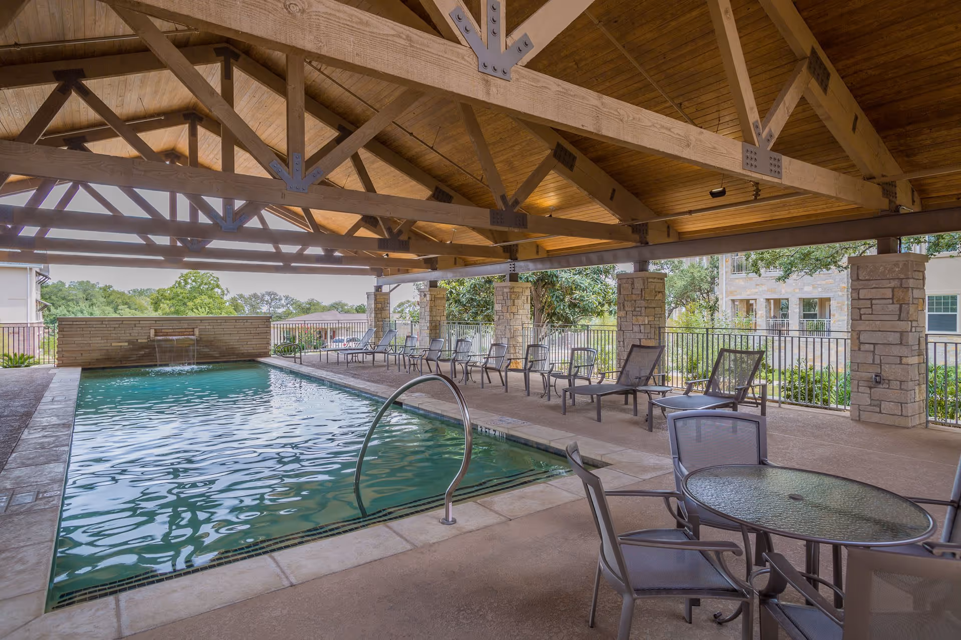 Covered outdoor swimming pool with a wooden beam ceiling, surrounded by lounge chairs and a glass-top table with chairs. Stone pillars support the roof and greenery is visible outside the fenced area.