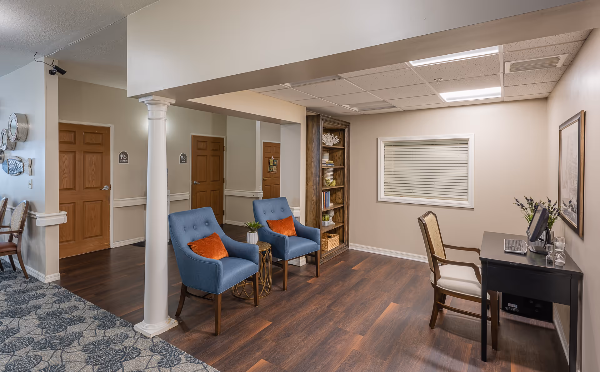 Interior view of a senior living facility showing a small seating area with two blue armchairs with orange cushions and a small round table between them. There is a wooden bookshelf with decorative items and books, a desk with a computer and a chair, and a window with closed blinds. The floor is wood with a patterned carpet in the adjacent area, and there are wooden doors and a white column in the background.