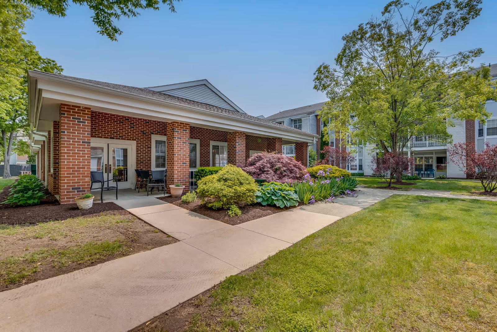 Exterior view of a senior living facility with a brick building featuring a covered patio area with chairs and tables. The surrounding area includes well-maintained landscaping with bushes, flowers, trees, and a concrete walkway leading to the entrance. Other residential buildings are visible in the background under a clear blue sky.