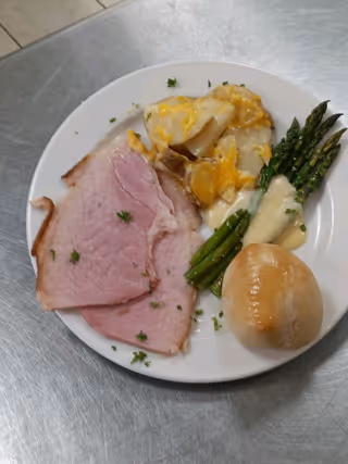 A white plate with two slices of cooked ham garnished with chopped herbs, a serving of scalloped potatoes topped with melted cheese, steamed asparagus with a creamy sauce, and a small round bread roll, all placed on a stainless steel surface.