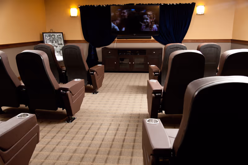 A small theater room with two rows of brown reclining chairs facing a wall-mounted flat screen TV. The room has beige walls with wooden trim, carpeted floor, and two wall sconces providing warm lighting. There is a framed photo collage on a small table to the left of the TV.