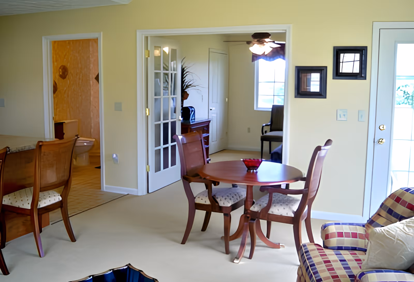 Bright residential living/dining area with a round wooden table and chairs, an upholstered armchair, and French doors leading to another room.