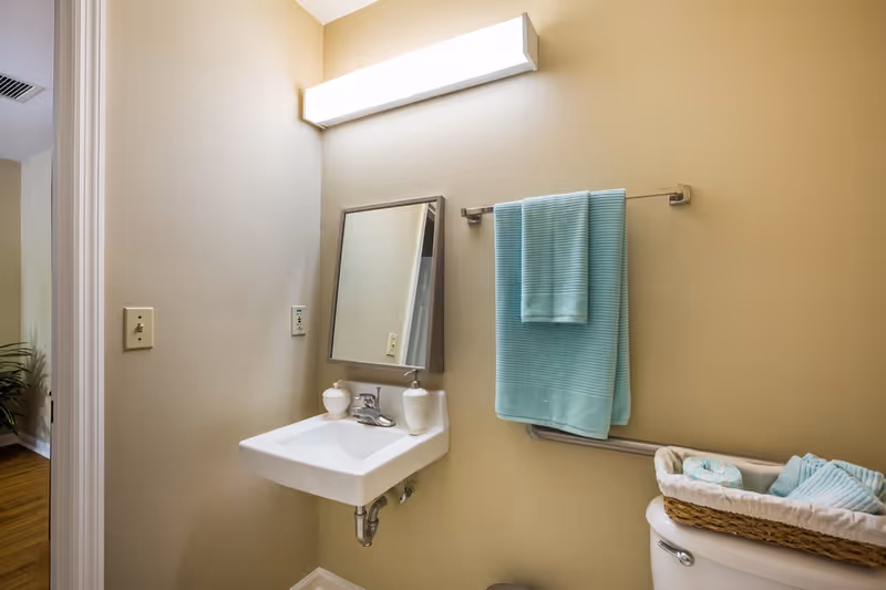 Small beige bathroom with a wall-mounted sink, angled mirror, teal towels, and a toilet topped with a woven basket of linens.