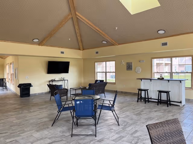 A spacious common area with a high vaulted ceiling and large windows letting in natural light. The room features a small round table with four blue chairs, a wicker seating area with a loveseat and two chairs around a coffee table, a wall-mounted flat-screen TV, and a white wooden bar with three black stools. The floor is tiled with light-colored tiles, and the walls are painted a soft yellow.