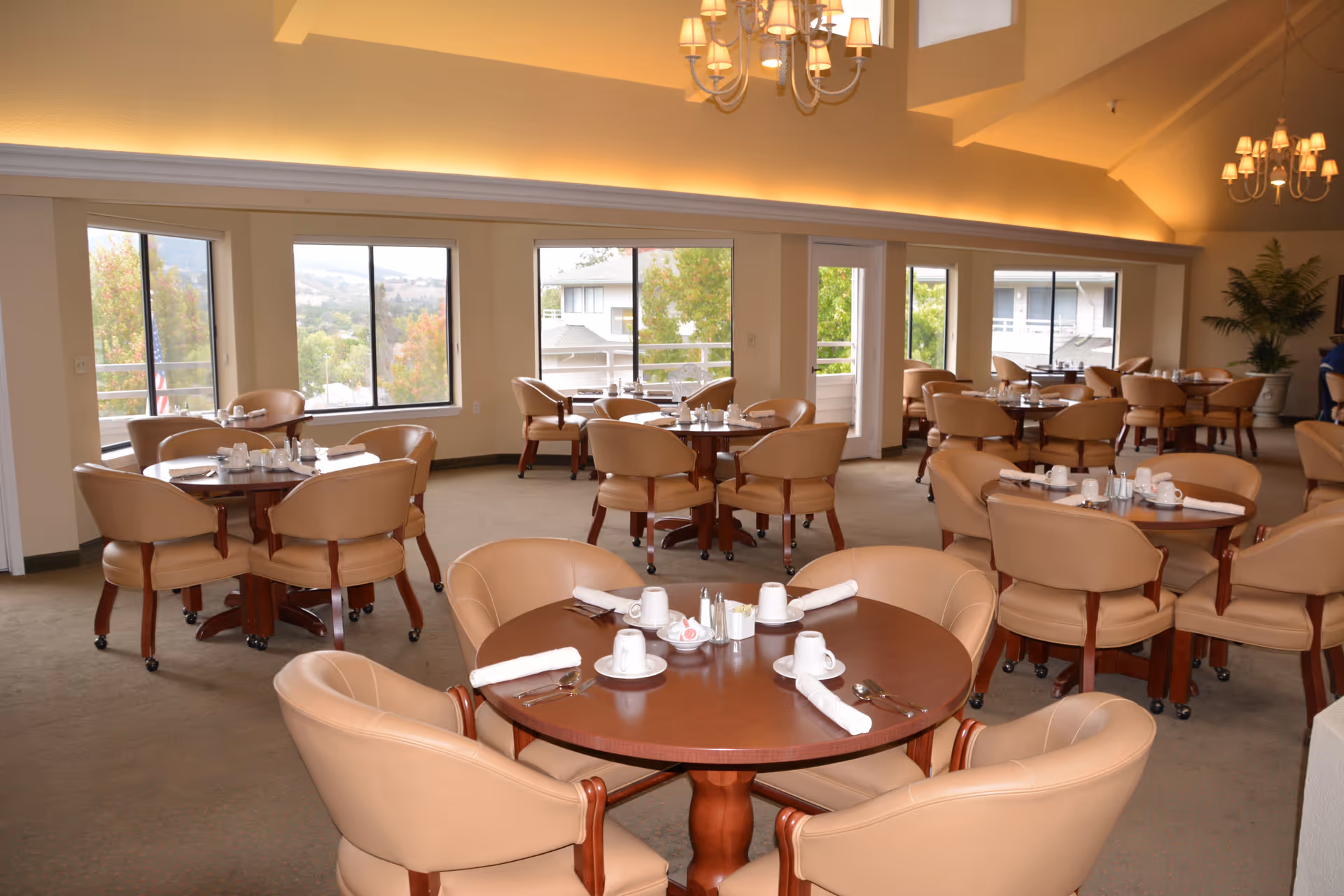 A spacious dining room with multiple round wooden tables, each surrounded by four beige cushioned chairs. The tables are set with white cups, saucers, rolled napkins, and silverware. Large windows line the walls, allowing natural light to brighten the room and providing views of trees and nearby buildings. The ceiling features chandeliers and recessed lighting, creating a warm and inviting atmosphere.