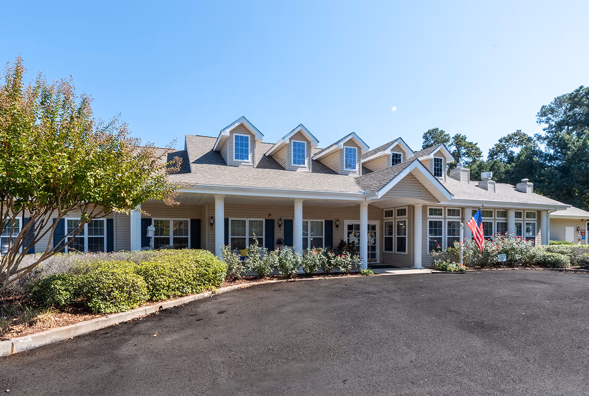 Exterior front view of a single-story senior living facility building with beige siding, multiple dormer windows on the roof, white columns supporting a covered entrance, and landscaped bushes and trees along the front. An American flag is displayed near the entrance under a clear blue sky.