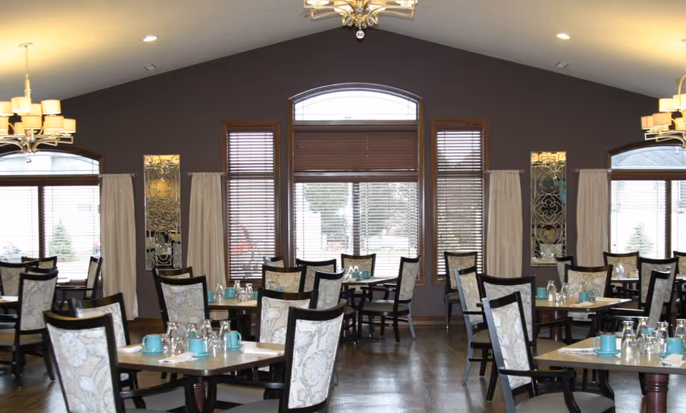 Dining room with multiple tables and chairs arranged neatly. Each table has turquoise cups, glasses, and napkins. The room has large windows with wooden blinds and curtains, dark purple walls, and two chandeliers hanging from the ceiling.