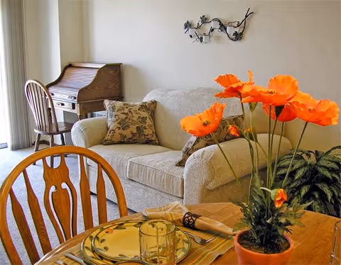 A cozy living room area with a beige sofa adorned with patterned cushions, a wooden roll-top desk with a chair in the corner, and a dining table in the foreground set with plates, a glass, and napkins. A vibrant orange flower arrangement in a pot is placed on the dining table, and a decorative metal wall art hangs above the sofa.