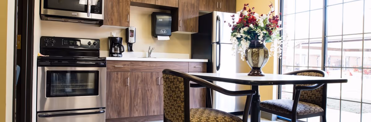 A kitchen area with stainless steel appliances including an oven and refrigerator, wooden cabinets, a coffee maker on the counter, and a dining table with two patterned chairs. A large window with grid panes lets in natural light, and a vase with red and white flowers is placed on the table.