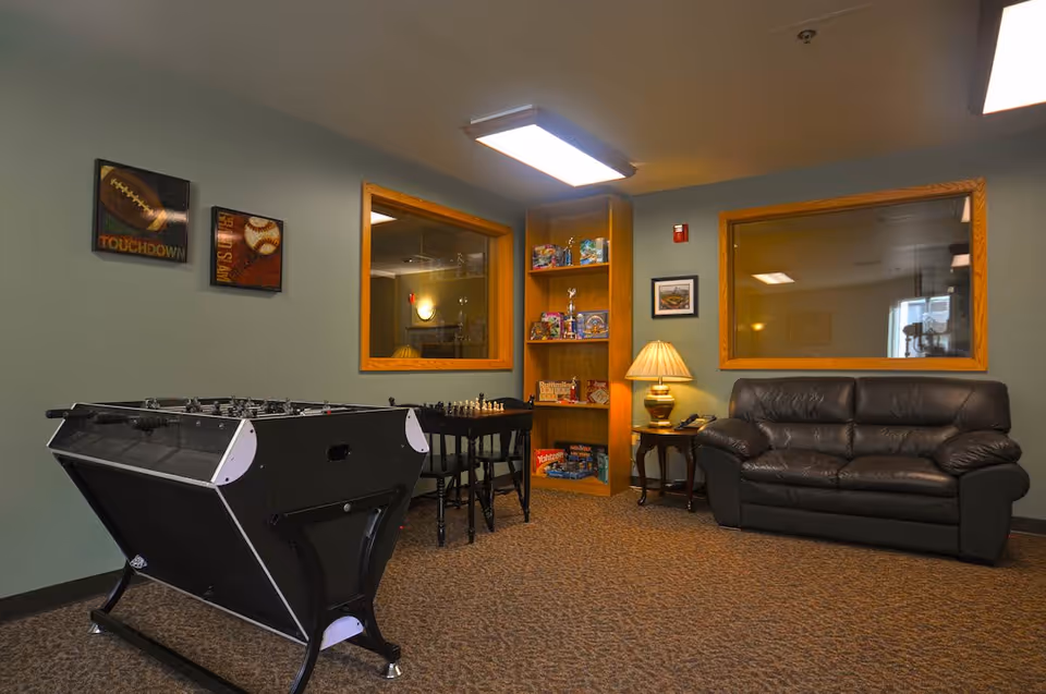 A recreational room with a black foosball table, a small table with chess pieces and chairs, a wooden bookshelf filled with board games and trophies, a side table with a lamp and telephone, and a black leather couch. The walls are painted green with two large windows and two sports-themed pictures hanging on the wall.
