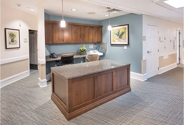 Interior view of a senior living facility reception or nurse station area with a large wooden counter in the foreground, a desk with office chairs, cabinets, a telephone, a sink, and framed floral artwork on the walls. The space has carpeted flooring and soft lighting.
