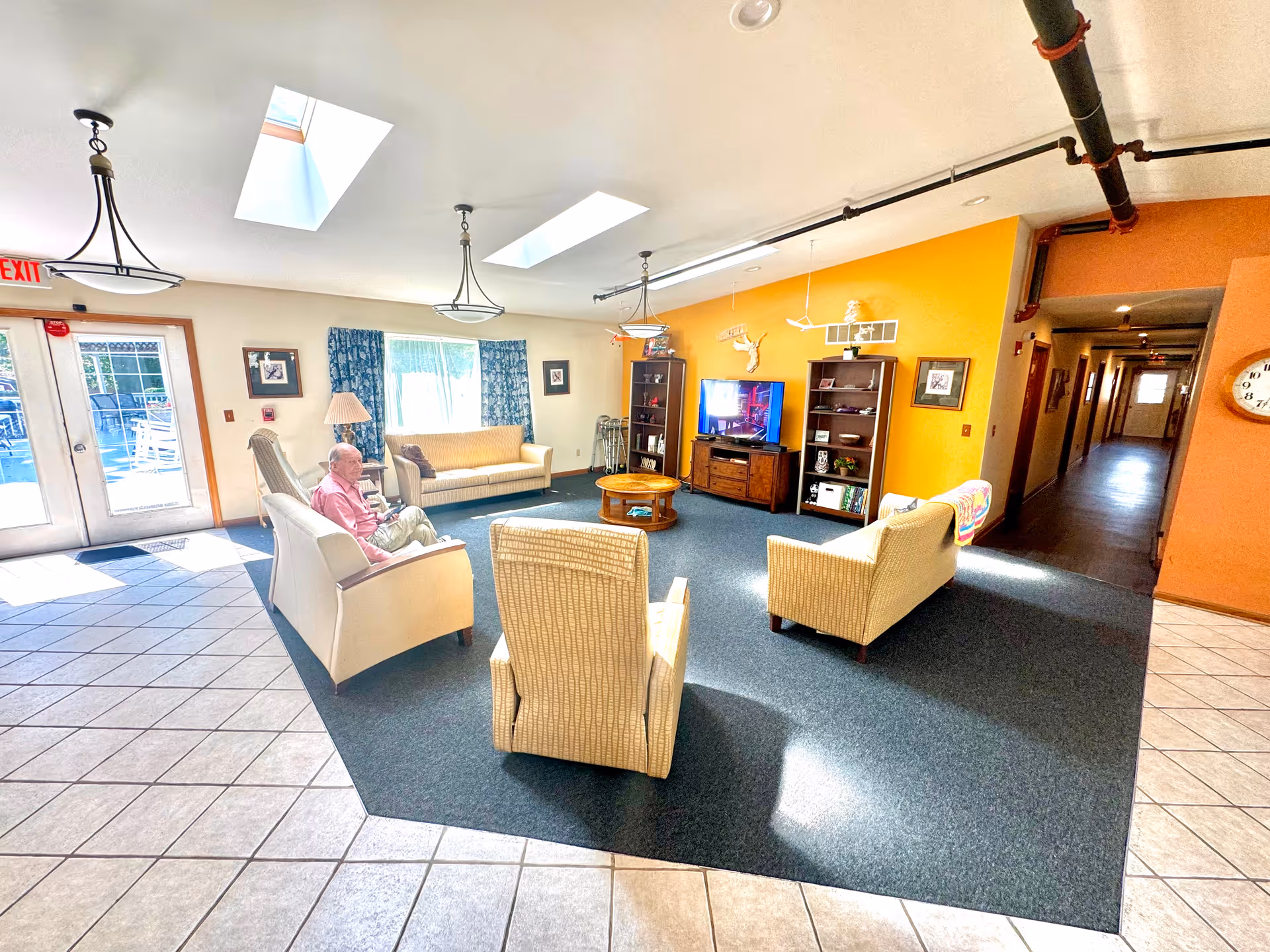 Bright communal lounge with sofas, armchairs, a TV, bookshelves and skylights in a senior living facility.