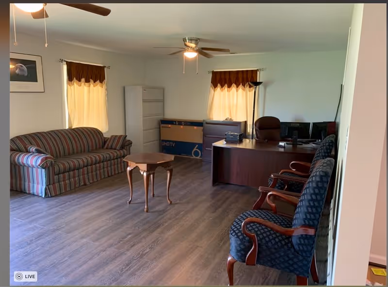 A room with wooden flooring featuring a striped couch on the left, a small wooden table in the center, and a dark wooden desk with office equipment on the right. There are two windows with yellow curtains, a ceiling fan with lights, and two upholstered chairs with wooden armrests in the foreground.