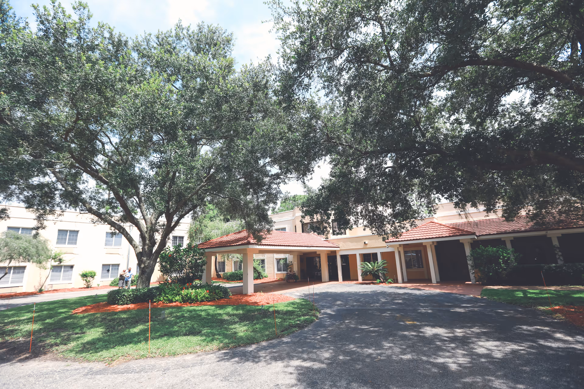 Exterior view of a senior living facility with a covered entrance, surrounded by large trees and landscaped greenery under a partly cloudy sky.