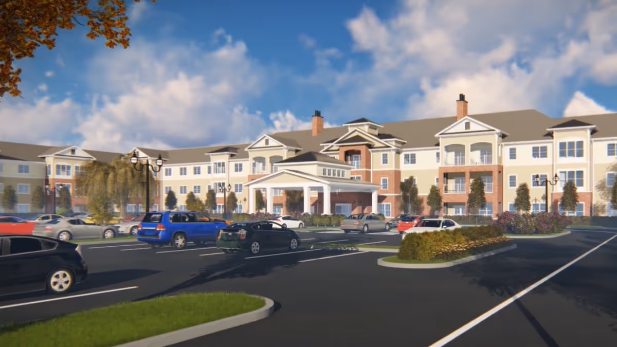 Exterior view of a large senior living facility building with multiple floors, balconies, and a covered entrance. Several cars are parked in the parking lot in front of the building under a partly cloudy sky.
