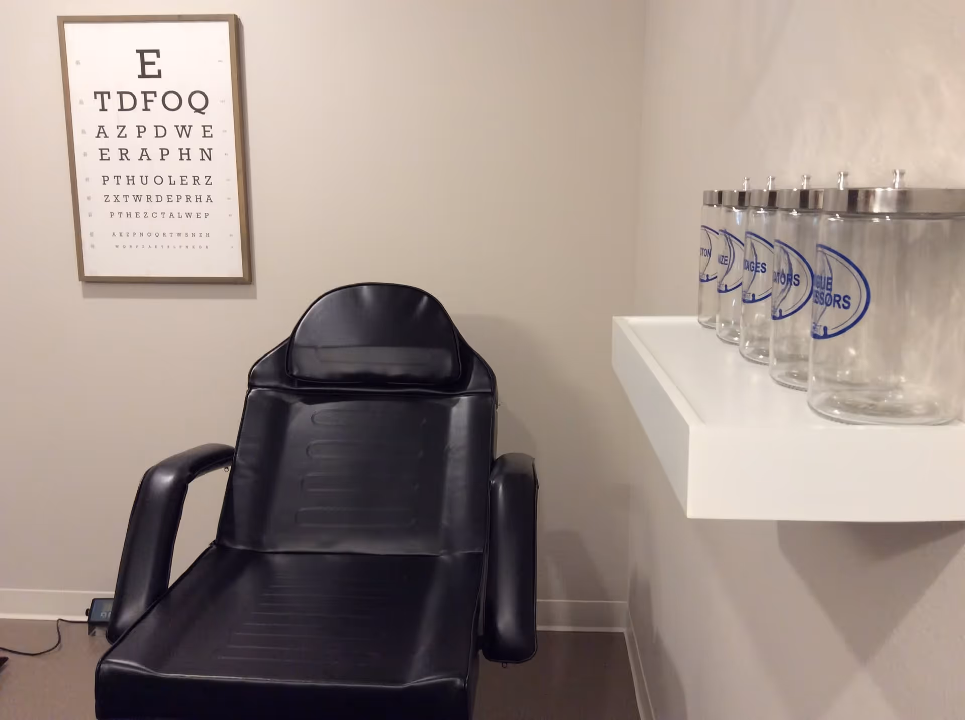 A small examination room with a black leather chair, an eye chart on the wall, and a white shelf holding several clear jars labeled with different types of eye accessories.