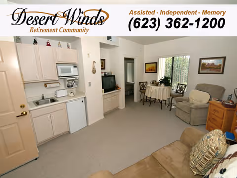 Interior view of a living area in Desert Winds Assisted Living facility showing a small kitchenette with cabinets, microwave, sink, and mini fridge on the left. The room has a carpeted floor, a round dining table with chairs near a window, a recliner chair, a wooden side table, and a couch with cushions. The walls are decorated with framed pictures.