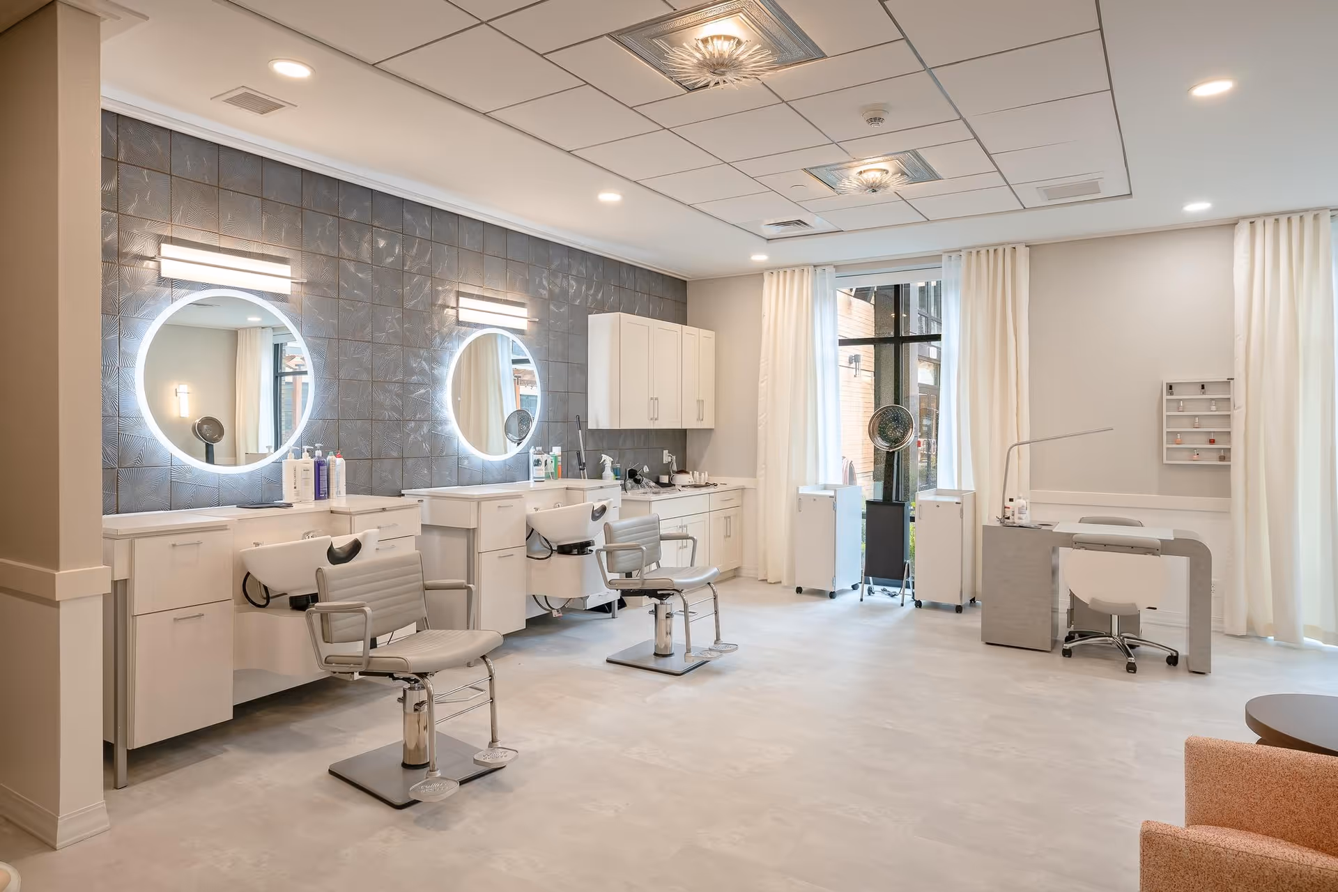 A bright and modern salon area with two hair washing stations and chairs in front of round mirrors with backlighting. The room has light-colored flooring, white cabinetry, and large windows with white curtains allowing natural light to enter. There is also a manicure station with a chair and nail polish display on the right side of the room.