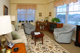 A cozy living room with two light blue tufted armchairs and a beige sofa with green cushions arranged around a wooden coffee table on a patterned rug. The room has large windows with beige valances, a wooden cabinet with decorative items, a vintage television, a table lamp, and framed pictures on the walls.