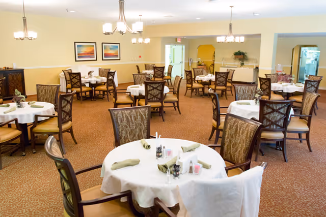 A spacious dining room with multiple round tables covered with white tablecloths and set with green napkins, salt and pepper shakers, and condiments. The room has beige walls, carpeted floors, several chandeliers, and framed artwork on the walls. Chairs with patterned upholstery surround each table.