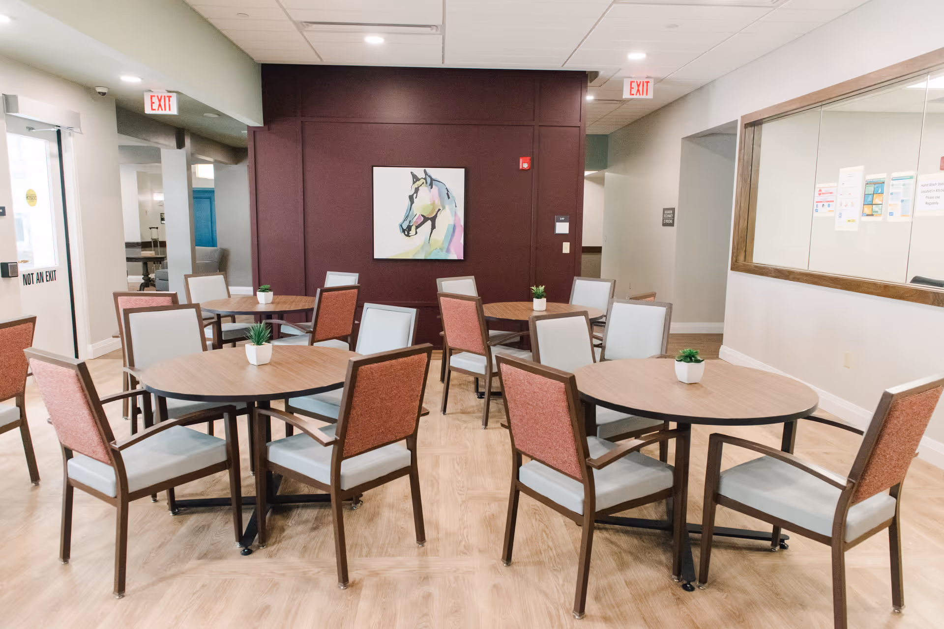 Communal dining room with round wooden tables, upholstered chairs, small potted plants, and a colorful horse painting on a burgundy accent wall.