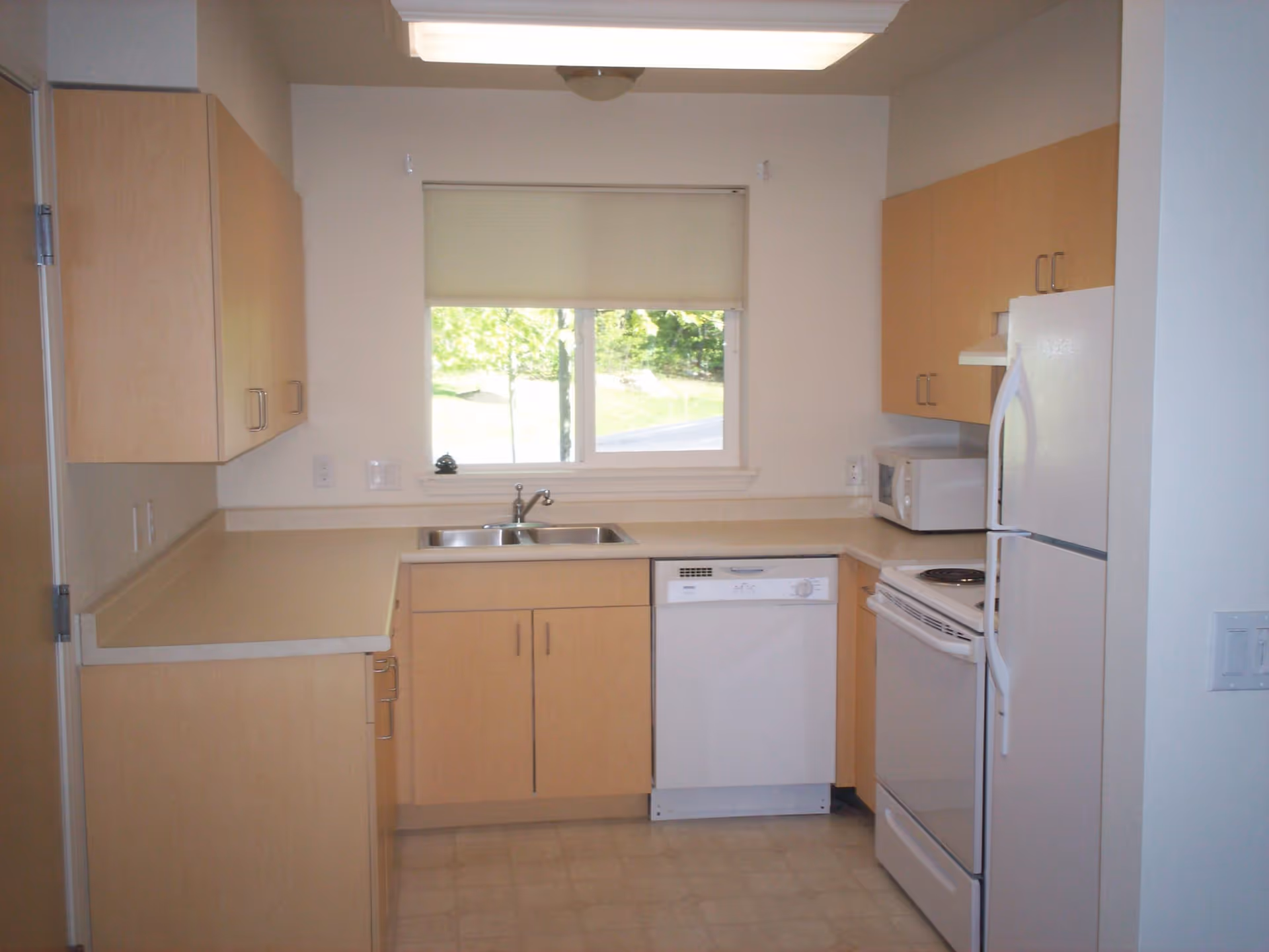 A clean, compact kitchen with light wood cabinets, a white refrigerator, white electric stove, white dishwasher, and a microwave on the counter. There is a double sink under a window with a beige roller shade, and the floor has a light-colored tile pattern.