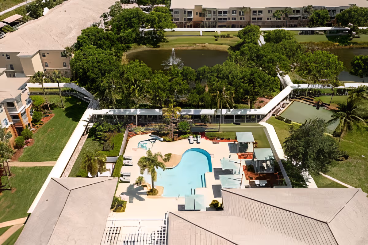 Aerial view of a senior living facility's outdoor area featuring a swimming pool surrounded by lounge chairs, palm trees, and landscaped greenery. In the background, there is a pond with a fountain and additional buildings and trees.