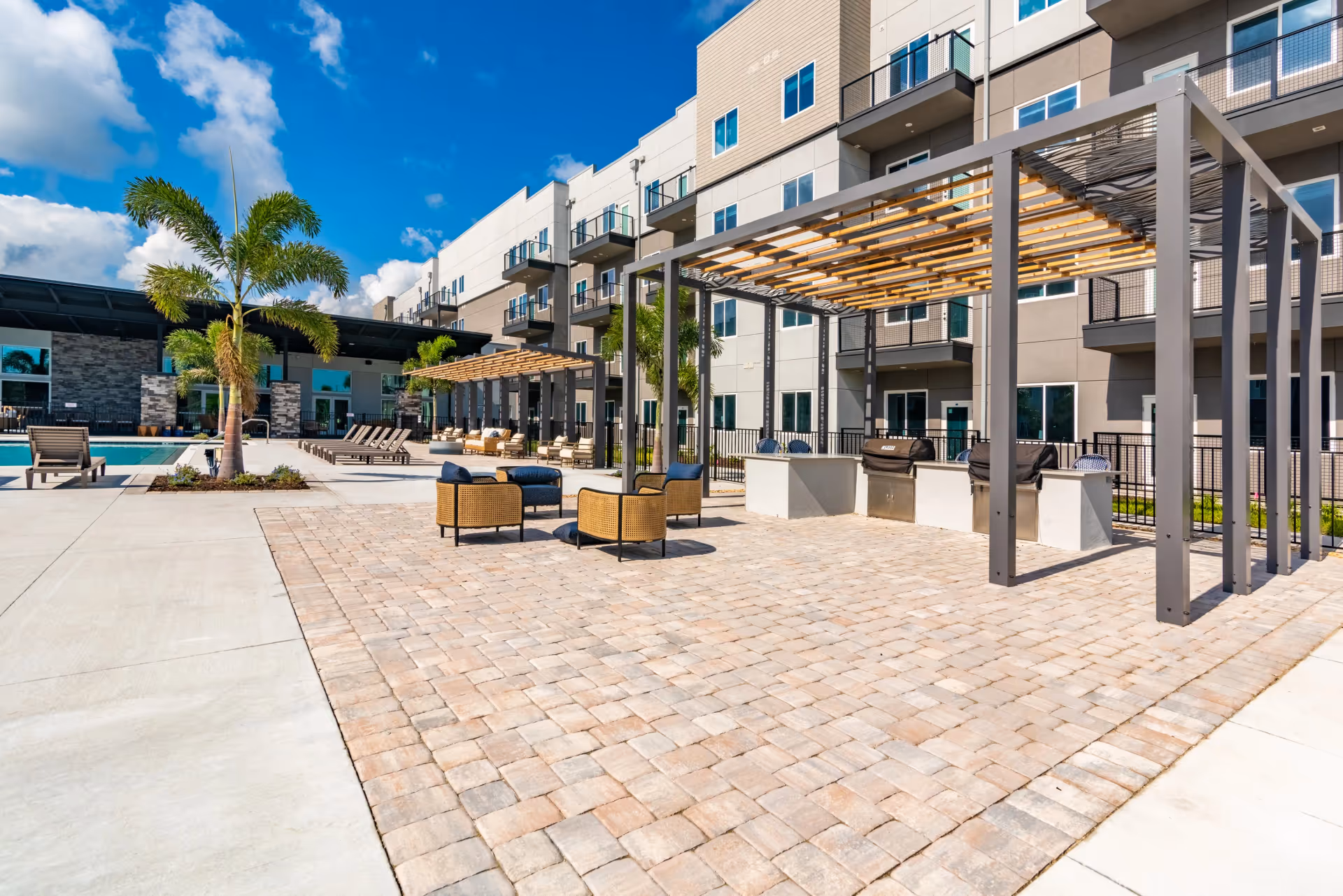 Outdoor courtyard and pool area with pergolas, lounge chairs, grills, and a multi-story building in the background.