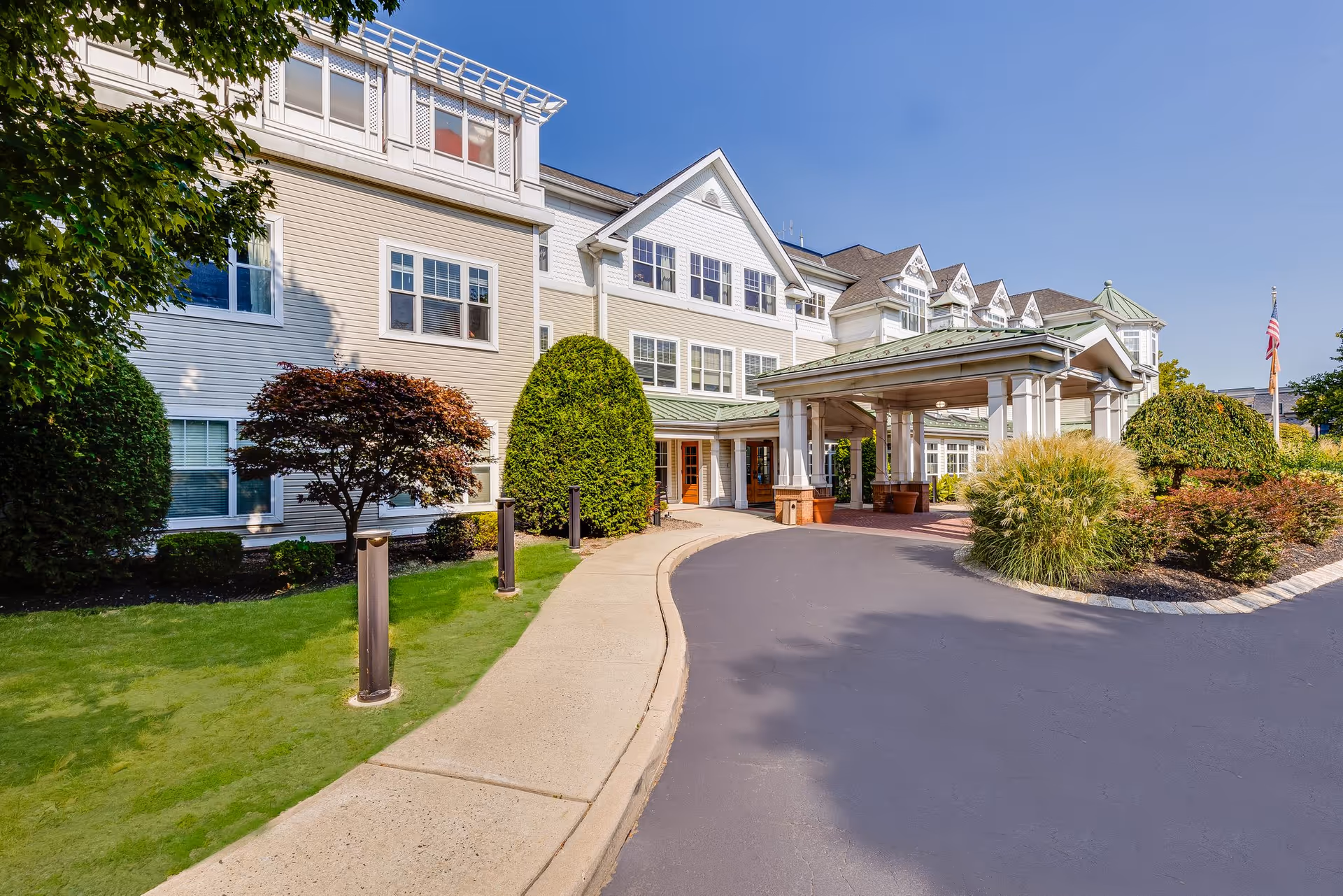 Exterior view of a senior living facility building with beige and white siding, multiple windows, and a covered entrance with a green roof. The surrounding area features well-maintained landscaping with bushes, trees, and a curved driveway leading to the entrance. An American flag is visible on the right side.