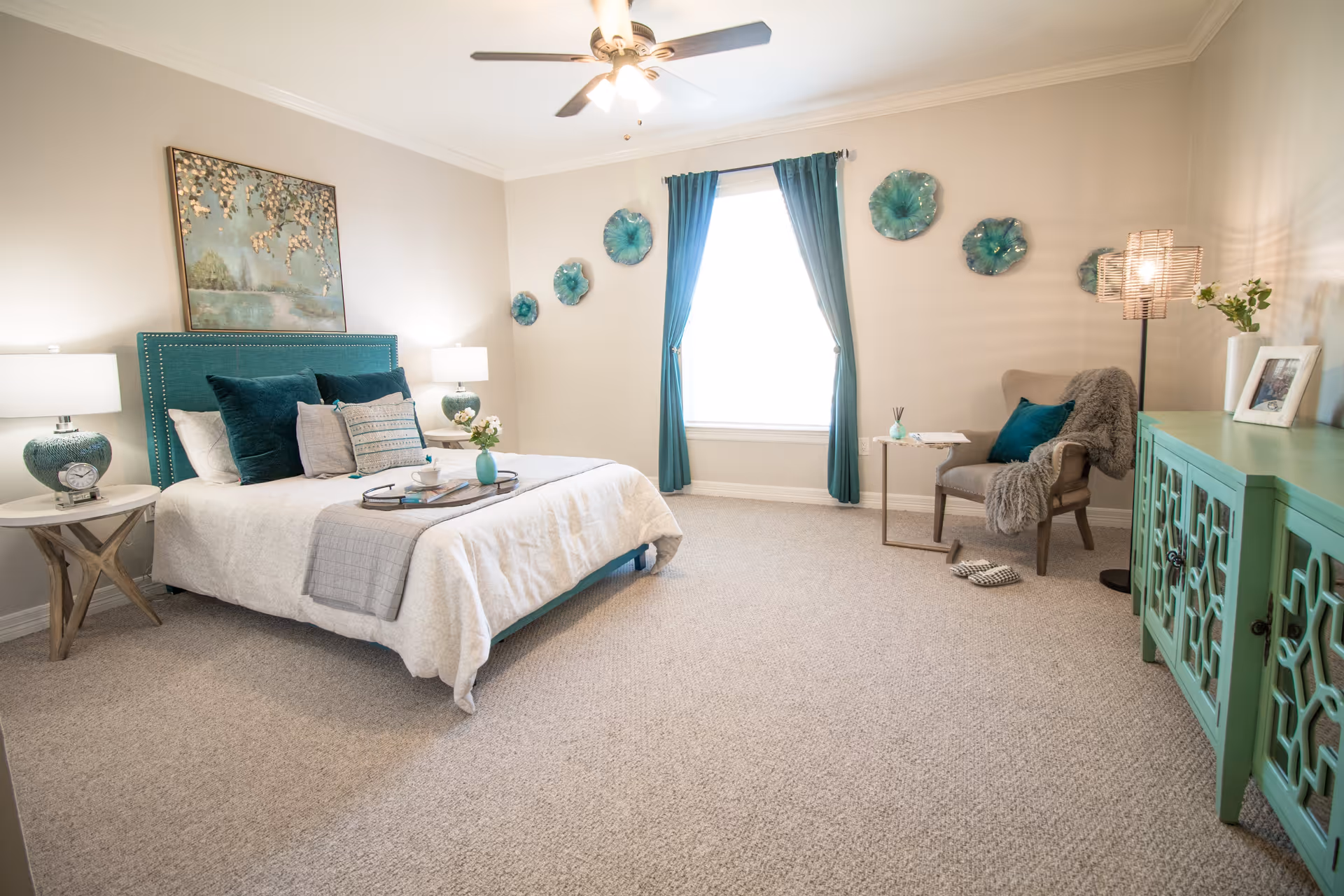 A bright and spacious bedroom with a large bed featuring teal and gray pillows and a teal headboard. There are two matching bedside tables with lamps, a ceiling fan with lights, teal curtains framing a window, decorative teal wall art, a comfortable armchair with a throw blanket and pillow, a small side table, and a teal cabinet with decorative details. The room has beige carpet and light-colored walls.