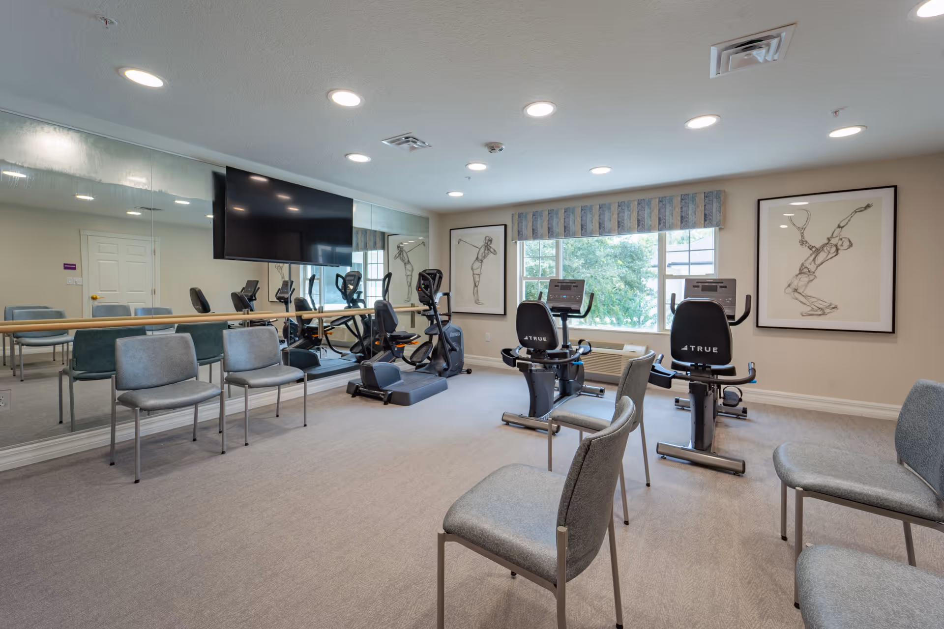Exercise room with two recumbent stationary bikes, several chairs, a wall-mounted TV, a full-length mirror, and framed artwork.