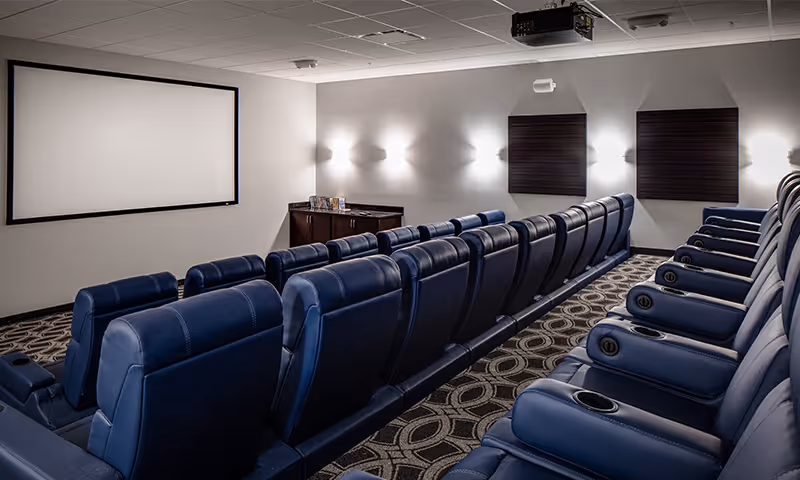A small theater room with two rows of blue leather recliner chairs facing a large blank projection screen on the wall. The room has patterned carpet, wall-mounted lights, and a projector mounted on the ceiling.