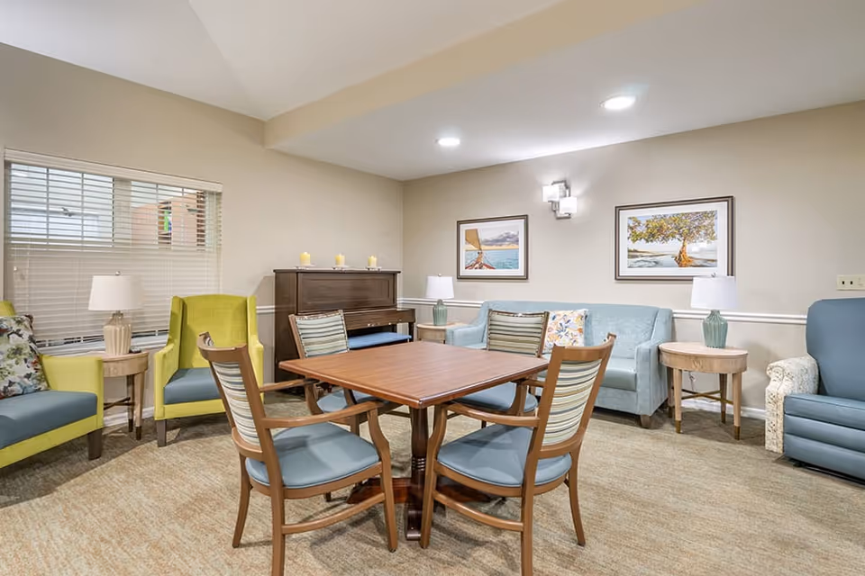 A cozy common area in a senior living facility featuring a square wooden table with four chairs in the center. Surrounding the table are various seating options including a light blue sofa, a green armchair, a blue armchair, and a patterned armchair. The walls are beige with two framed landscape paintings and two table lamps on wooden side tables. A piano with candles on top is placed against the back wall beneath a window with blinds.