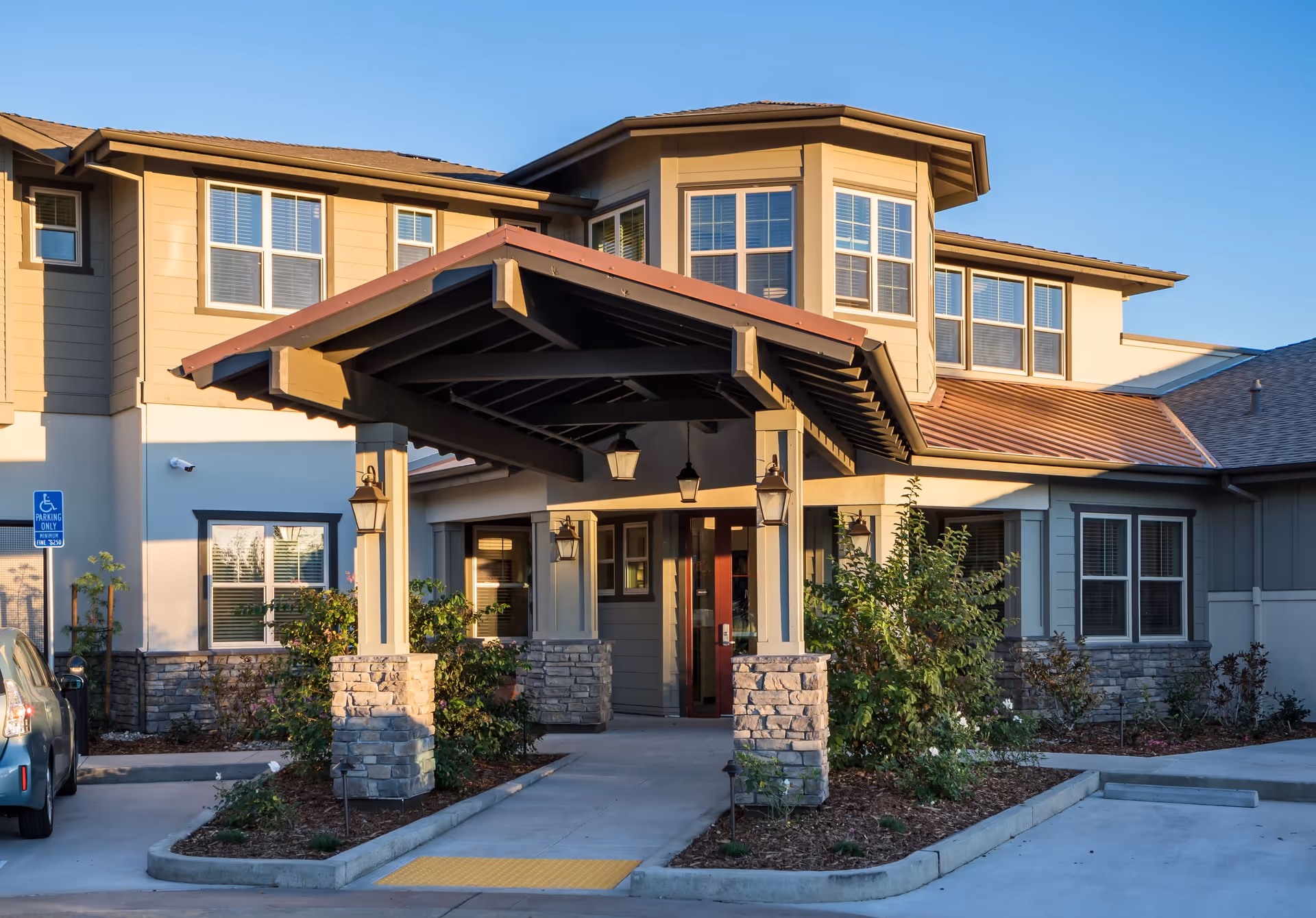 Exterior view of a senior living facility entrance with a covered drop-off area supported by stone and wooden pillars, surrounded by landscaping and a parking area with a handicapped parking sign.